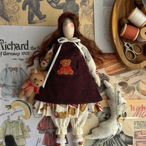 Puede incluir: Una muñeca de tela con cabello largo castaño que viste un vestido de peto de pana marrón oscuro sobre una camisa color crema con estampado de ositos. La muñeca sostiene un pequeño osito de peluche marrón con una cinta roja y usa bombachos.
