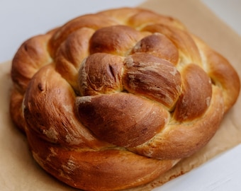 May include: A golden-brown, braided loaf of bread, possibly challah, sits on a sheet of parchment paper. The bread has a glossy crust and a detailed, woven pattern. The baked goods are a light brown color.