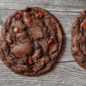 May include: Two chocolate chip cookies with a dark brown, almost black, color. The cookies have a slightly cracked surface and are covered in chocolate chips.