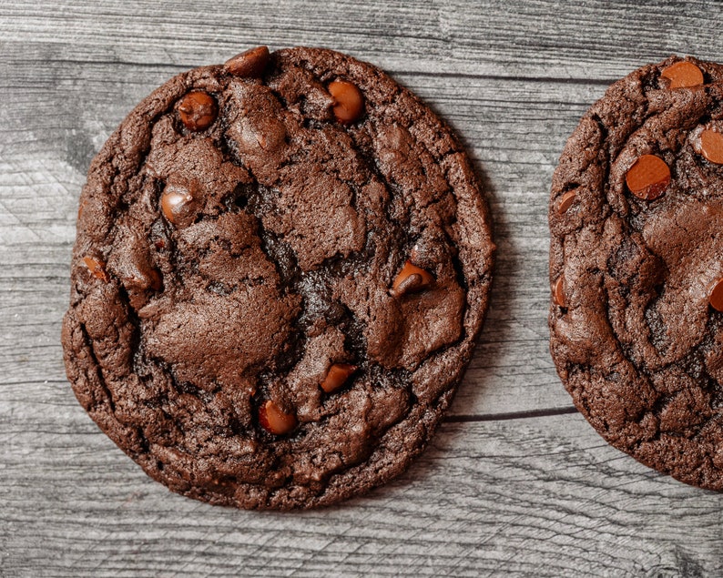 May include: Two chocolate chip cookies with a dark brown, almost black, color. The cookies have a slightly cracked surface and are covered in chocolate chips.