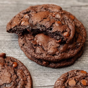May include: Three chocolate chip cookies on a wooden surface. The cookies are dark brown and have a slightly crispy texture. The cookies are arranged in a pile, with the top cookie slightly broken.