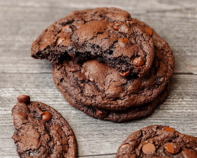 May include: Three chocolate chip cookies on a wooden surface. The cookies are dark brown and have a slightly crispy texture. The cookies are arranged in a pile, with the top cookie slightly broken.