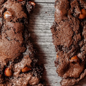 May include: Two broken pieces of a chocolate chip cookie on a wood background. The cookie is dark brown and has chocolate chips throughout.