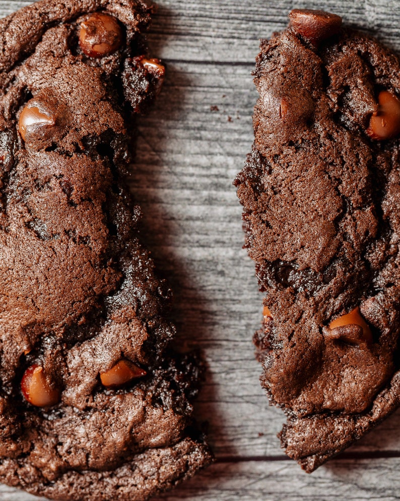 May include: Two broken pieces of a chocolate chip cookie on a wood background. The cookie is dark brown and has chocolate chips throughout.