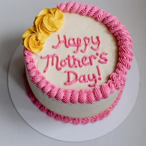 May include: A white cake with pink frosting and a pink and white swirl design around the edge. The cake is decorated with yellow flowers and the words "Happy Mother's Day!" in pink frosting.
