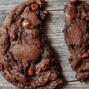 May include: A broken chocolate chip cookie with a dark brown color and visible chocolate chips. The cookie is on a wooden surface.
