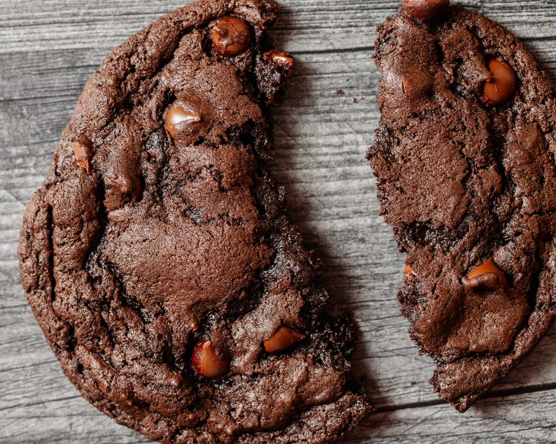 May include: A broken chocolate chip cookie with a dark brown color and visible chocolate chips. The cookie is on a wooden surface.