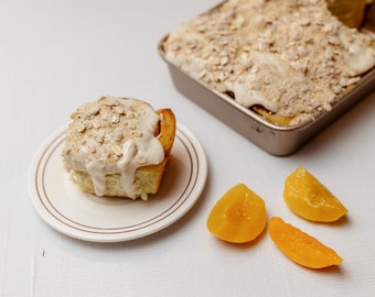 Op de afbeelding: Een enkele kaneelrol met witte glazuur en haverkrumble topping op een wit bord. Drie perzikplakjes liggen op een wit oppervlak naast het bord. Een bakvorm met meer kaneelrollen is op de achtergrond.