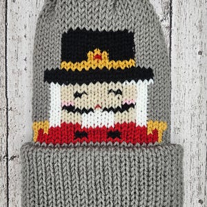 Puede incluir: Gorro de punto gris con un diseño de cascanueces de punto de colores en la parte delantera. El cascanueces tiene un sombrero negro, una chaqueta roja y una cara blanca.