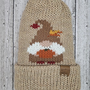 Può includere: Un berretto di lana beige con un design a gnomo marrone con un cappello rosso, una barba bianca, una zucca arancione e fiamme gialle. Il berretto ha un'etichetta in pelle marrone con il testo "KCH" su di essa.