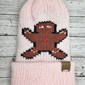 May include: Pink knitted beanie with a brown gingerbread man design and a brown leather patch with the text "KGY" on the side.
