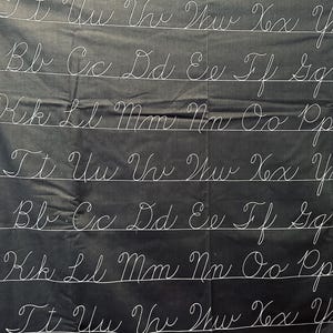 May include: Black fabric with white cursive letters, including the alphabet. The letters are arranged in rows with horizontal lines. The fabric has a slightly wrinkled texture.