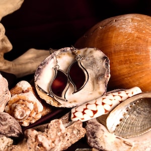 May include: A pair of silver dangle earrings with dark red glass teardrop-shaped stones. The earrings are displayed on a bed of seashells.