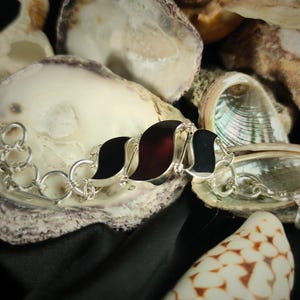 Könnte beinhalten: Ein silbernes Kettenarmband mit drei schwarzen und braunen ovalen Anhängern. Das Armband wird auf einem Bett aus Muscheln präsentiert.