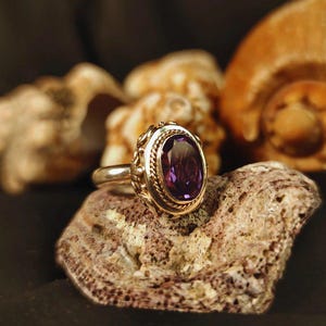 May include: A gold and silver ring with a large oval purple gemstone. The ring is set on a textured, light brown surface, with seashells in the background. The gemstone is a deep, rich purple color.