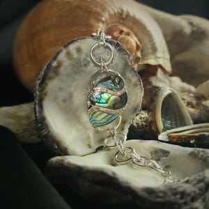 Könnte beinhalten: Ein silbernes Armband mit einem großen Abalone-Muschel-Anhänger. Der Anhänger ist schillernd in Grün-, Blau- und Violetttönen. Das Armband wird auf einem Muschel-Hintergrund präsentiert.