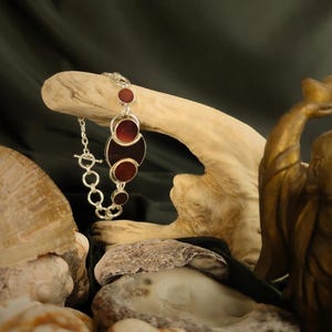 Könnte beinhalten: Ein silbernes Armband mit einem Kettenglieddesign und fünf roten und schwarzen runden Steinen. Das Armband wird auf einem Stück Treibholz präsentiert.