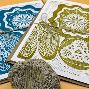 May include: Two handmade greeting cards with intricate white designs on a blue and green background. The cards are tied together with twine and are sitting on a brown surface.