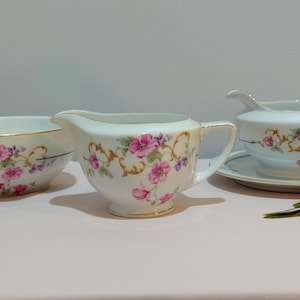 May include: A vintage porcelain tea set featuring a bowl, creamer, and gravy boat. Each piece is white with gold trim and adorned with pink and purple floral patterns. The set is displayed on a light pink surface.