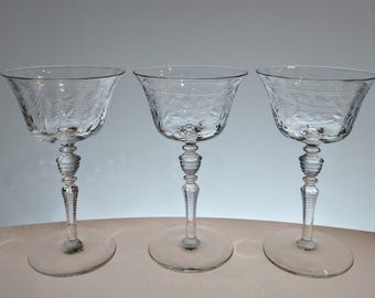 May include: Three clear, stemmed glasses with etched floral designs. The glasses have a rounded bowl, a decorative stem, and a circular base. The glasses are arranged in a row, showcasing their elegant design.