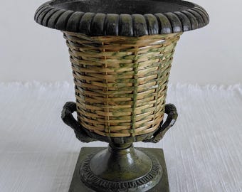 May include: A decorative urn featuring a woven wicker body with green and tan accents, set within a dark metal frame. The piece includes a wide, fluted rim, two small side handles, and a square base, all finished in a dark, weathered metal.