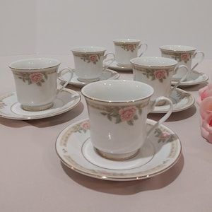 May include: A set of six white teacups and saucers. Each cup and saucer features a delicate pink rose design with gold trim. The cups have a classic shape, perfect for serving tea or coffee.