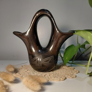 May include: A dark brown, handcrafted vase with a unique handle and a decorative etched design. The vase has a wide opening and a rounded base. The vase is sitting on a beige doily.