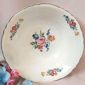 May include: A round, cream-colored ceramic bowl with a scalloped edge and a metallic gold rim. The interior features a central floral bouquet of pink, yellow, and blue flowers, with smaller matching floral clusters placed along the inner rim.