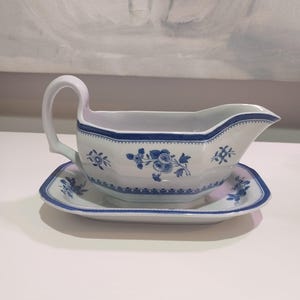 May include: White porcelain gravy boat and matching saucer with blue floral patterns and trim. The gravy boat has a curved handle and a spout for easy pouring. The saucer is rectangular with rounded corners. A classic addition to any table setting.