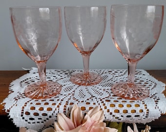 May include: Three pink-tinted glass goblets with a delicate etched pattern. The goblets have a stemmed base and a wide bowl. They are arranged on a white lace doily, with pink flowers in the foreground. The goblets are likely vintage or antique.