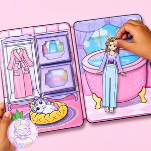 May include: A paper doll playset featuring a bathroom and closet scene. The set includes a paper doll character, a pink bathtub, a purple wardrobe with a pink robe, and a small unicorn in a yellow bed. The scene is illustrated in pastel colors.