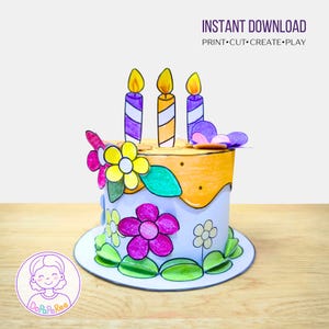 Op de afbeelding: Een papieren verjaardagstaart met drie kaarsen en kleurrijke bloemen. De taart heeft een witte basis met oranje glazuur en is versierd met paarse, gele en roze bloemen. De tekst "INSTANT DOWNLOAD" staat bovenaan.