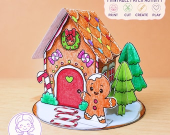 May include: A 3D paper craft gingerbread house with a red door, a green wreath, and colorful candy decorations. A smiling gingerbread person stands in front, accompanied by two green paper trees on a circular base. Text reads PRINTABLE PAPER ACTIVITY.