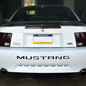 Puede incluir: Un Ford Mustang Mach 1 blanco con luces traseras oscurecidas y una calcomanía negra "MUSTANG" en el parachoques trasero. El coche tiene una matrícula de Pensilvania.