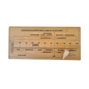 May include: A wooden Bovis scale with text in Spanish, including "Radiaciones de los seres humanos" and "Luz Ultravioleta." A crystal pendulum rests on the scale. The scale measures various levels, including "Cancer" and "Saludable."