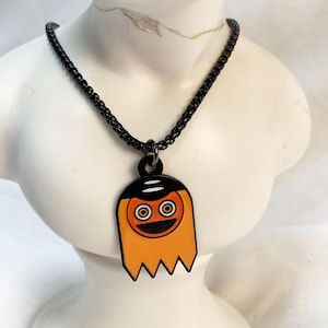 Op de afbeelding: Een zwarte metalen ketting met een hanger in de vorm van een gestileerde geest. De geest heeft een zwart bovendeel, een oranje lichaam en een rond oranje gezicht met grote ogen en een zwarte mond. De hanger hangt aan een donkere schakelketting.