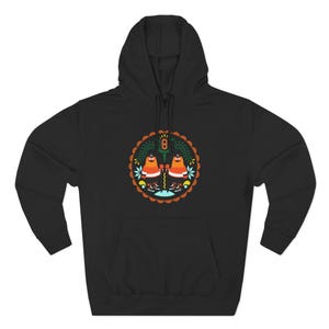 May include: A black hooded sweatshirt featuring a central circular graphic. The design displays two orange, bird-like figures with white accents, surrounded by stylized green foliage, light blue floral elements, and a scalloped brown border.