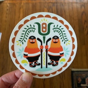 Könnte beinhalten: Runder Aufkleber mit zwei Cartoon-Charakteren in orangefarbenen und schwarzen Hockey-Uniformen, die Hockeyschläger halten. Der Aufkleber hat einen weißen Hintergrund mit einem dekorativen Rand und floralen Elementen. Die Charaktere haben orangefarbene Körper, schwarze Streifen und schwarze Schlittschuhe.