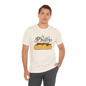 Könnte beinhalten: Ein cremefarbenes Kurzarm-T-Shirt mit einer grafischen Illustration eines Cheesesteak-Sandwichs. Über dem Sandwich steht das Wort Philly in einer stilisierten, dampfenden Schriftart. Das Design verwendet braune, gelbe und schwarze Farben.