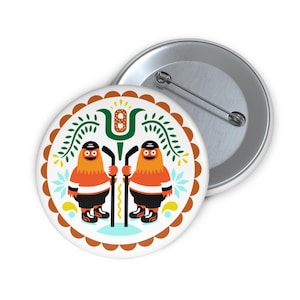 Könnte beinhalten: Ein runder Ansteckbutton mit einer grafischen Illustration von zwei orangefarbenen, pelzigen Wesen in schwarzer Eishockeyausrüstung, die Eishockeyschläger halten. Das Design zeigt ein zentrales grünes Blumenmotiv, einen braunen Wellenrand und hellblaue Akzente.
