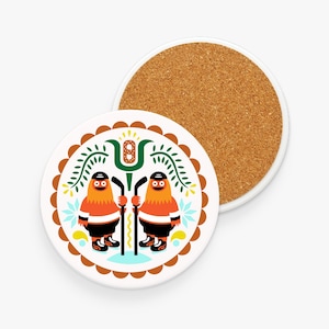 Könnte beinhalten: Zwei runde Untersetzer. Einer zeigt ein Cartoon-Design von zwei orangefarbenen Figuren in Hockeyausrüstung, die Hockeyschläger halten. Das Design umfasst grüne Blätter, blaue Blumen und einen braunen, gewellten Rand. Der zweite Untersetzer ist aus Kork.