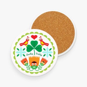 Könnte beinhalten: Zwei runde Untersetzer. Einer hat eine weiße Keramikoberfläche mit einem grün-weißen Blumendesign, roten Vögeln und dem Text "Lucky Gritty". Der andere Untersetzer hat eine Korkoberfläche.