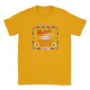 Può includere: Una maglietta gialla con una grafica in stile vintage di un prodotto di panini di patate chiamato "Martin's 8 Sandwich Potato Rolls". La grafica presenta un bordo rosso, giallo e verde con un motivo a cuore rosso.