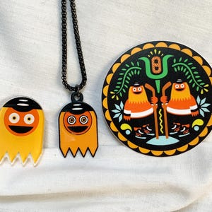 Puede incluir: Un conjunto que incluye un pin de acrílico con forma de fantasma amarillo, un colgante de metal a juego con forma de fantasma en una cadena negra y una pegatina circular con dos figuras de fantasmas naranjas con palos de hockey.