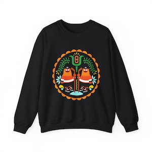 Op de afbeelding: Zwart sweatshirt met een kleurrijk cirkelvormig ontwerp met twee oranje figuren die hockeysticks vasthouden, omringd door groene bladeren en oranje en blauwe accenten.