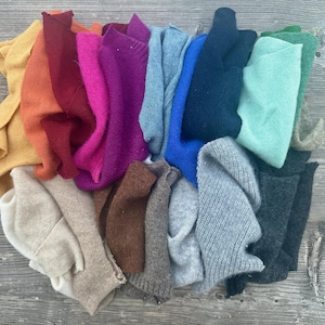 Könnte beinhalten: Eine Sammlung gefalteter Pullover in verschiedenen Farben, darunter Gelb, Orange, Rot, Pink, Lila, Blau, Türkis, Grau und Braun. Die Pullover sind auf einer verwitterten Holzoberfläche angeordnet und zeigen eine lebendige Farbpalette.
