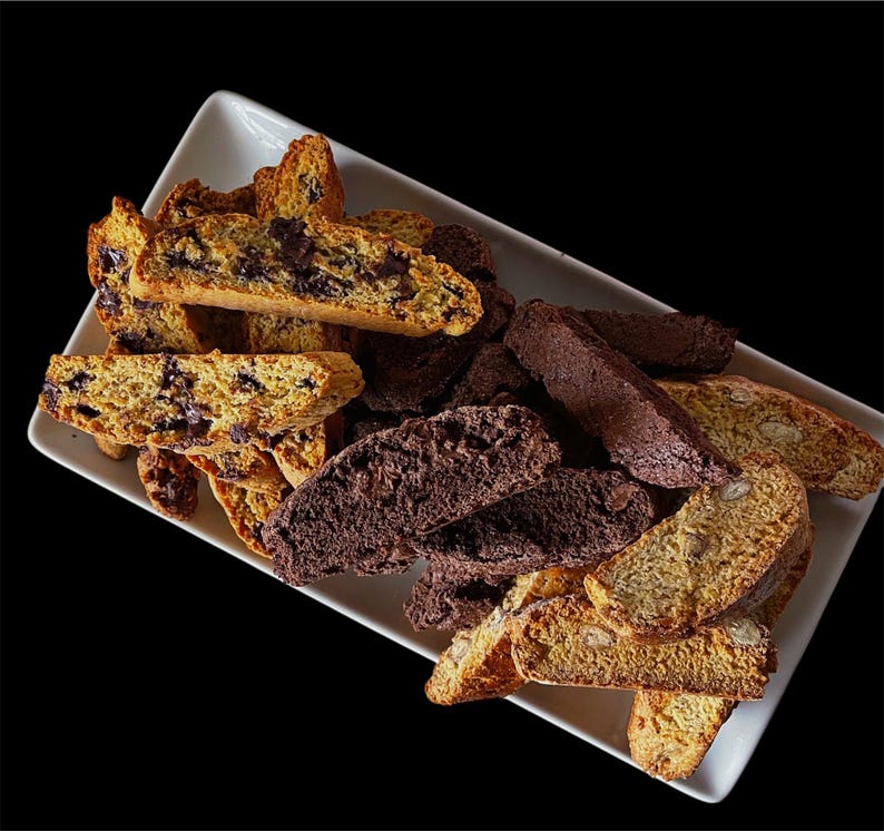 May include: A white rectangular plate filled with a variety of biscotti. The biscotti are both chocolate and plain. Some of the biscotti are broken into pieces. The biscotti are arranged in a random pattern on the plate.