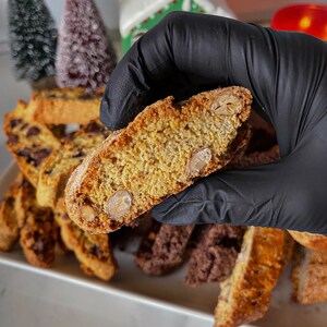 May include: A close-up of a biscotti cookie broken in half, revealing the almond pieces inside. The cookie is light brown in color and has a slightly crispy texture. The cookie is surrounded by other biscotti cookies, some of which are chocolate flavored.
