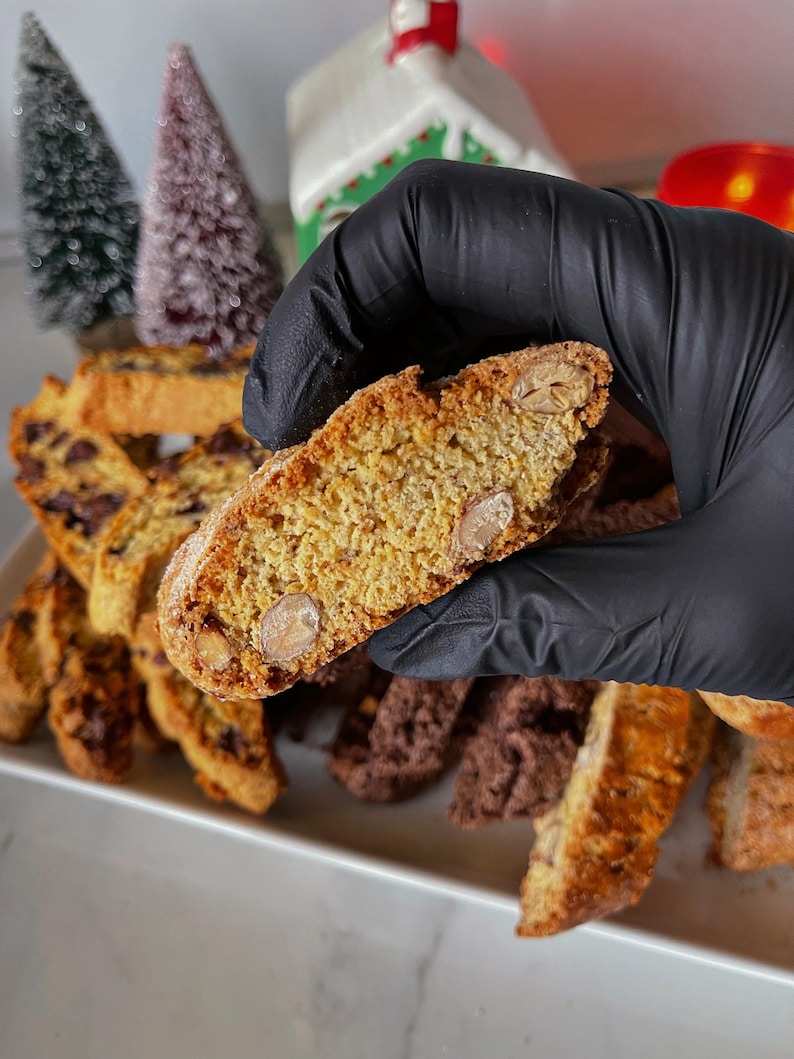 May include: A close-up of a biscotti cookie broken in half, revealing the almond pieces inside. The cookie is light brown in color and has a slightly crispy texture. The cookie is surrounded by other biscotti cookies, some of which are chocolate flavored.