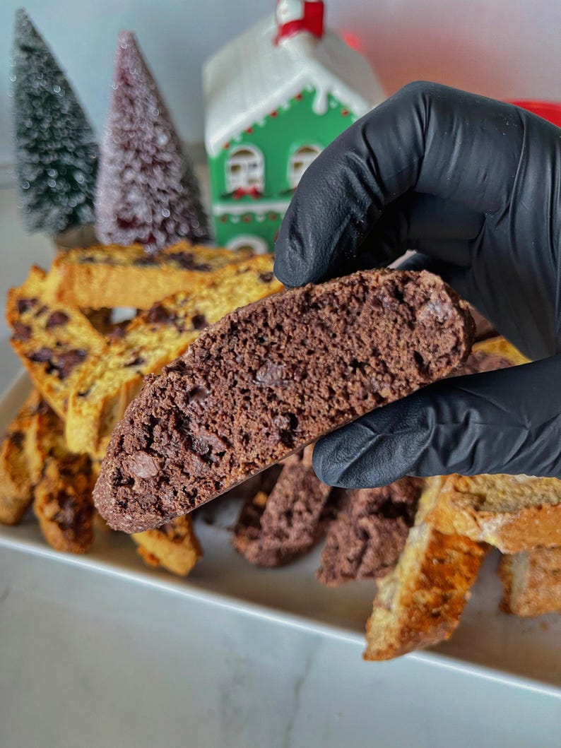 May include: A close-up of a chocolate biscotti being held by a gloved hand. The biscotti is broken in half, revealing the chocolate chips inside. There are other biscotti in the background, some are chocolate and some are plain.